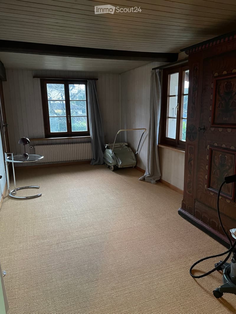 Carpeted room with wooden walls, large windows with curtains, a small round table with a lamp, and a radiator beneath the windows.