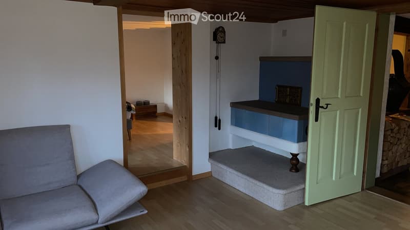 A cozy living room with a grey sofa, a wooden floor, a small staircase leading to a raised platform, and a clock on the wall. The room is connected to another space with a green door.