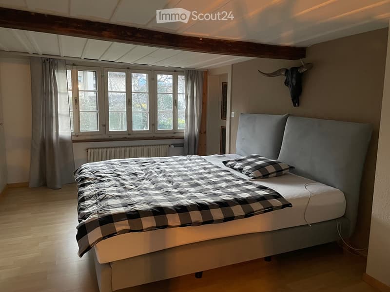 Cozy bedroom with wooden flooring, large windows with curtains, a grey upholstered bed with a checkered duvet, and a decorative wall piece.