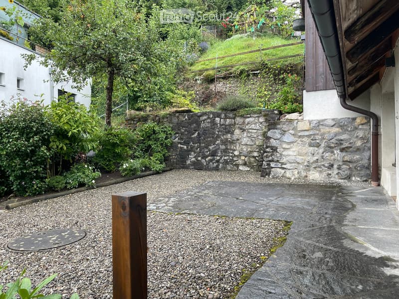 Garden area with stone walls, gravel, and a wooden post, surrounded by greenery and a raised section of grass.