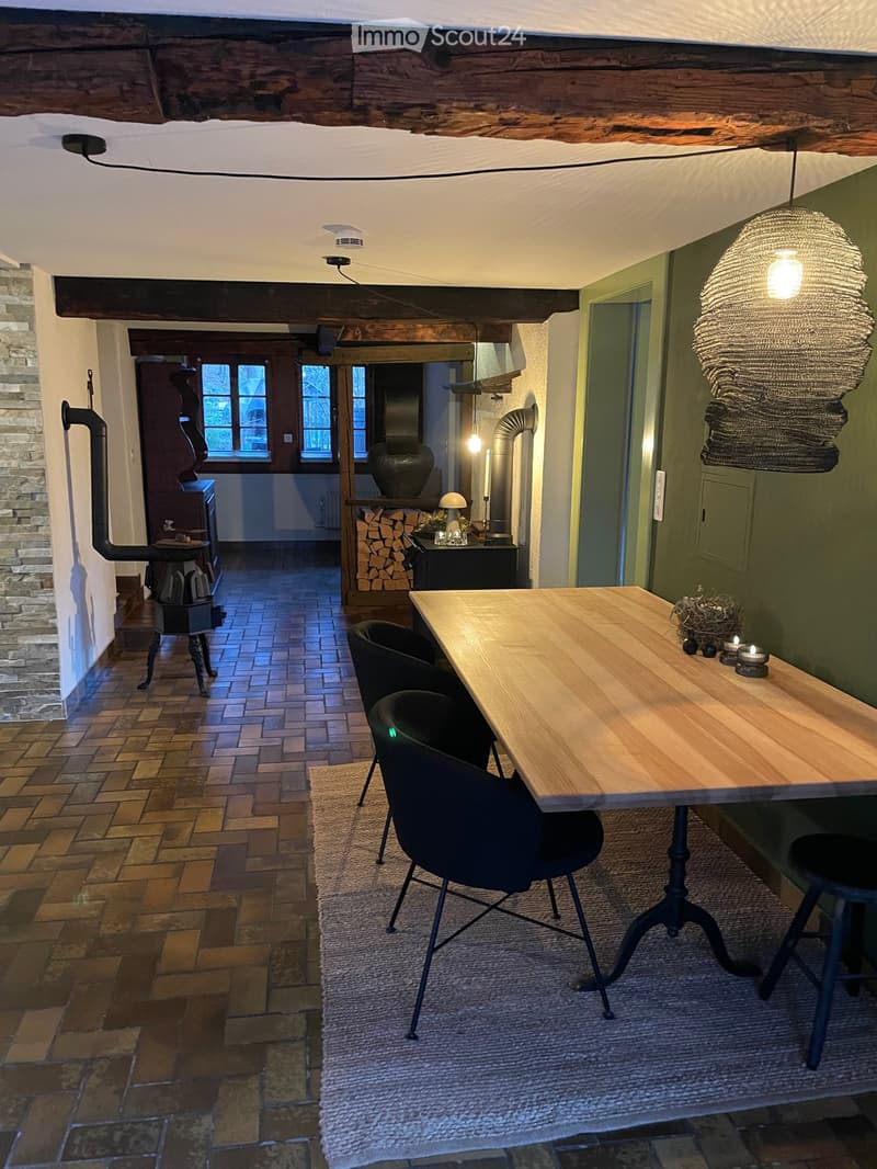 Cozy dining area with a wooden table, chairs, and a stool, featuring a wood stove, a vase, and a decorative lamp.