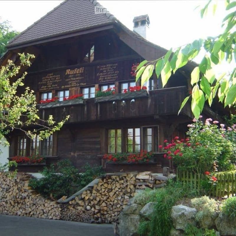 Traditional wooden house with a steep roof, multiple windows, and flower boxes on the balconies. The house is surrounded by greenery and a stack of firewood is visible in front.