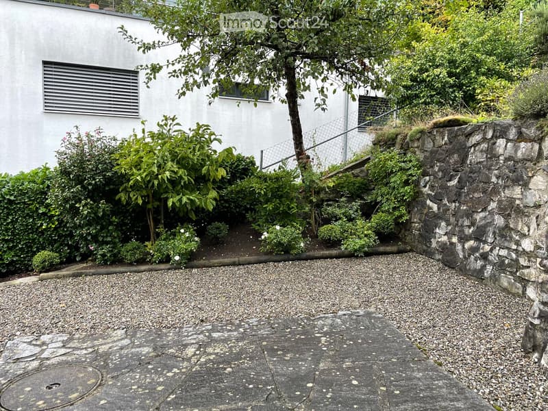 Garden area with gravel and stone pathways, surrounded by greenery and a white building with windows and a metal fence.