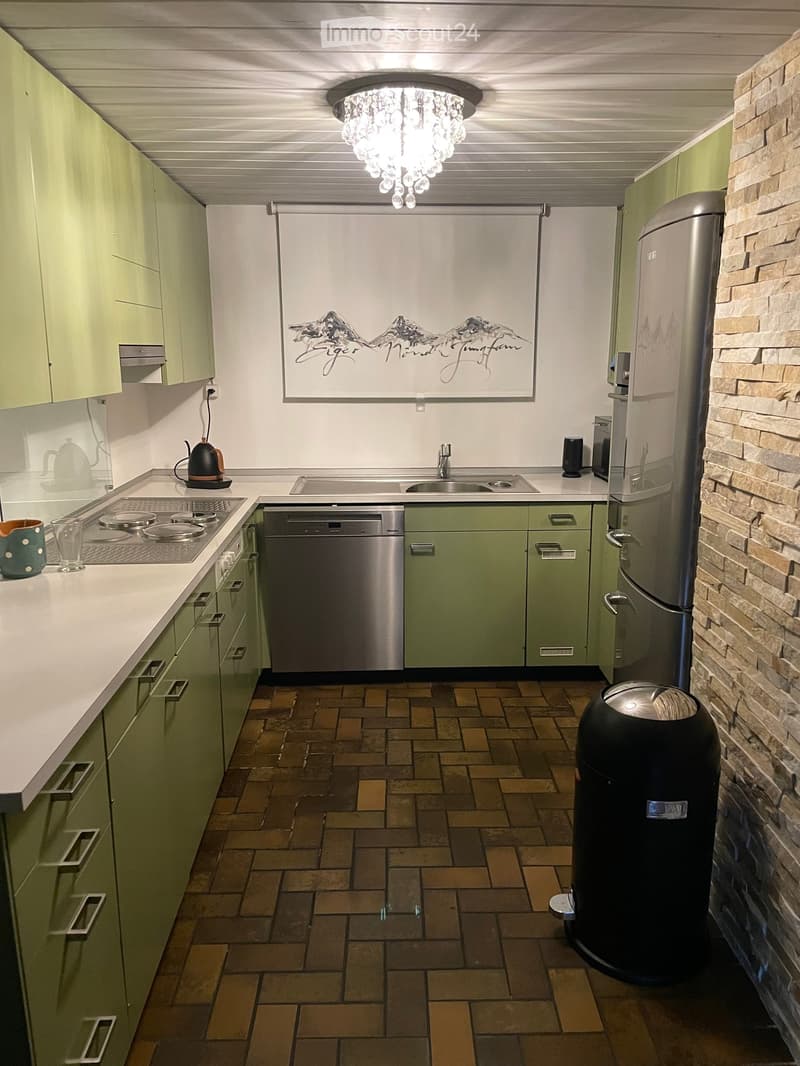 Modern kitchen with green cabinets, a central island, stainless steel appliances, and a brick accent wall. Features a chandelier light fixture and a framed mountain artwork above the sink.