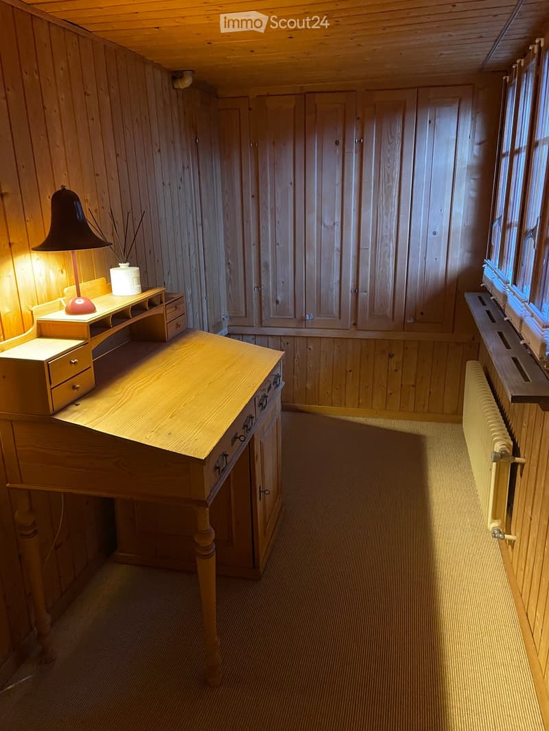 A small wooden-paneled room with a large wooden table, a lamp on a small wooden stand, a radiator, and built-in wooden cabinets.