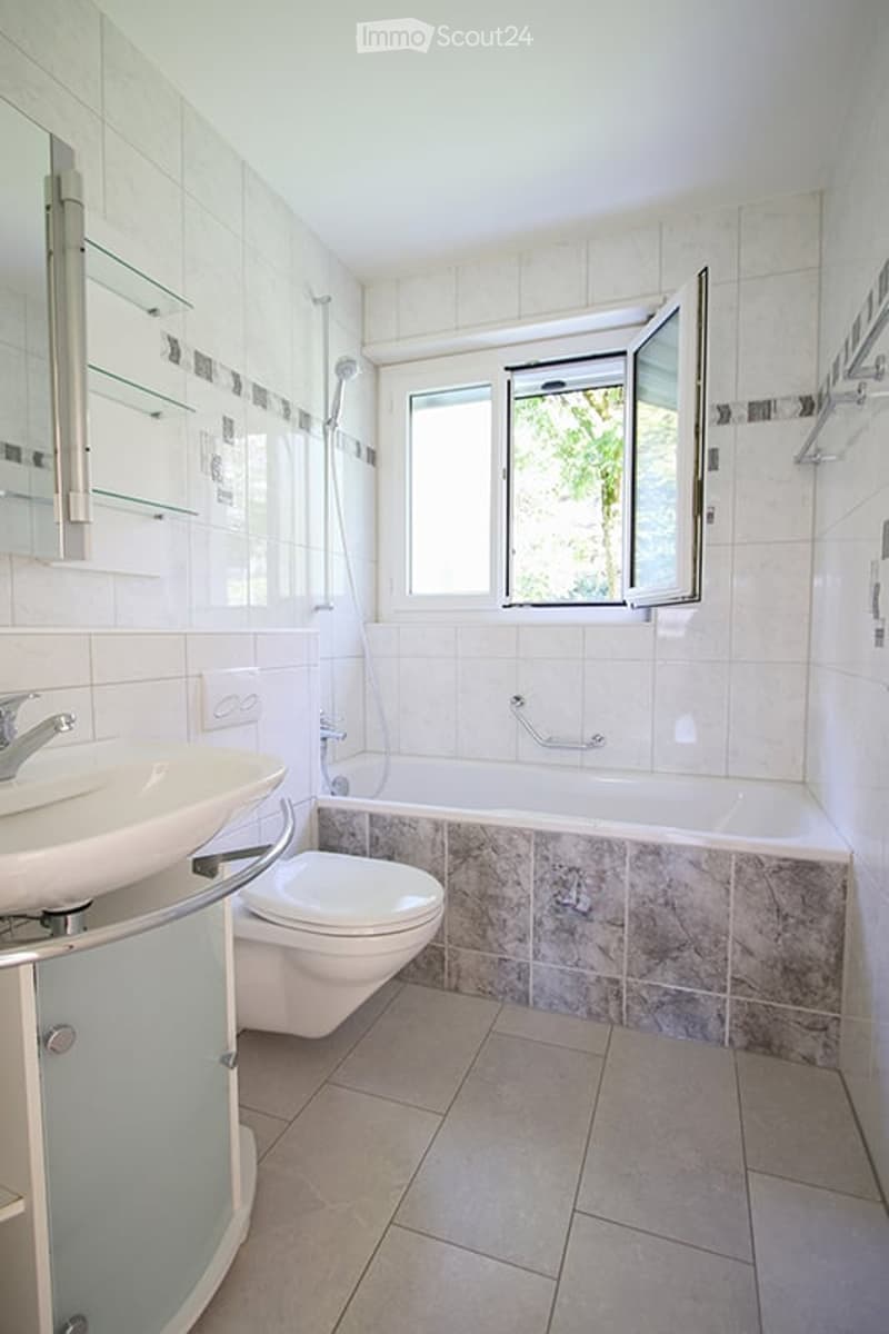 Modern bathroom with white tiles, a sink, toilet, bathtub, and a large window with blinds.