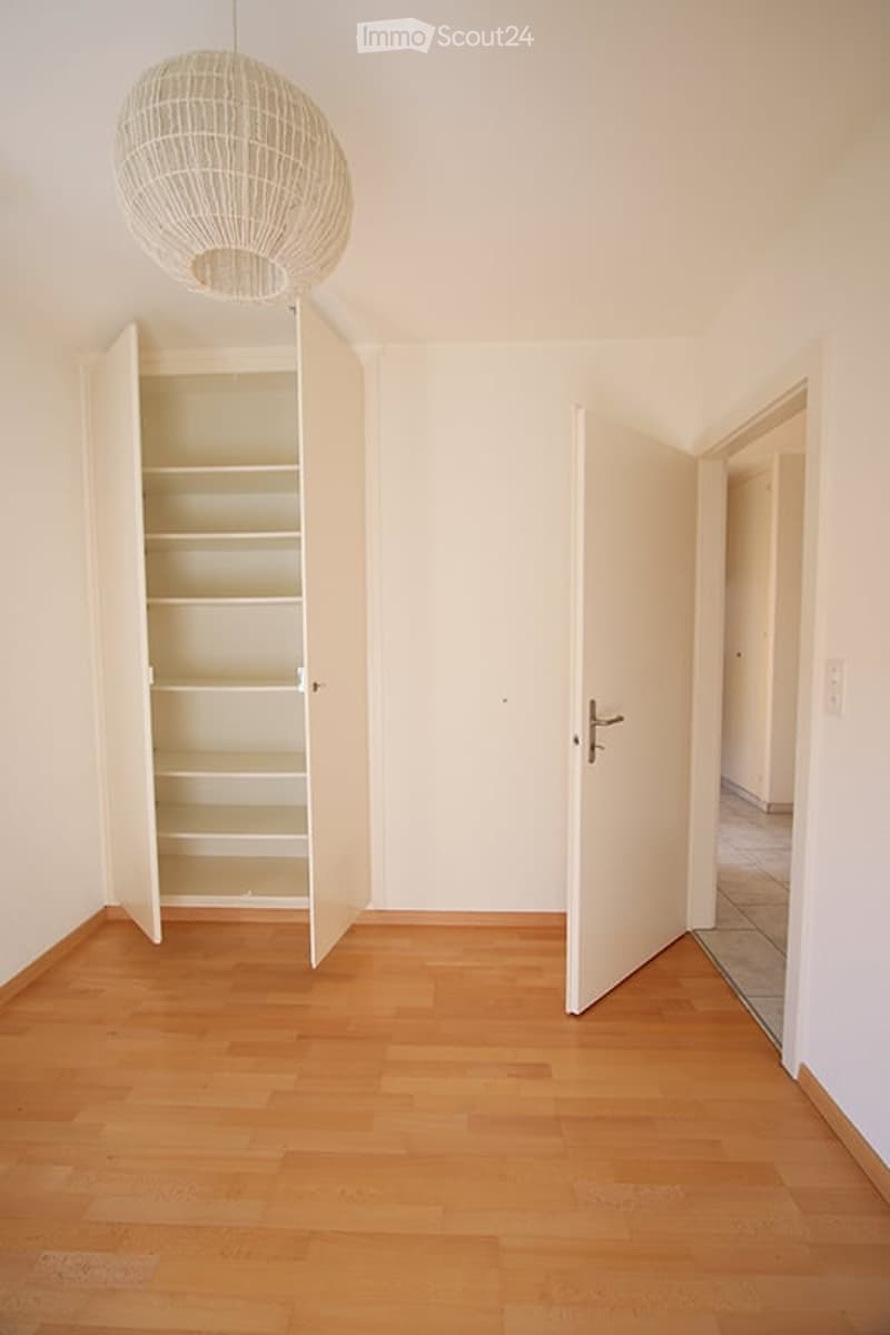 Completely empty room with light wooden flooring, white walls, and a built-in closet with open shelves. A large, round pendant light hangs from the ceiling.