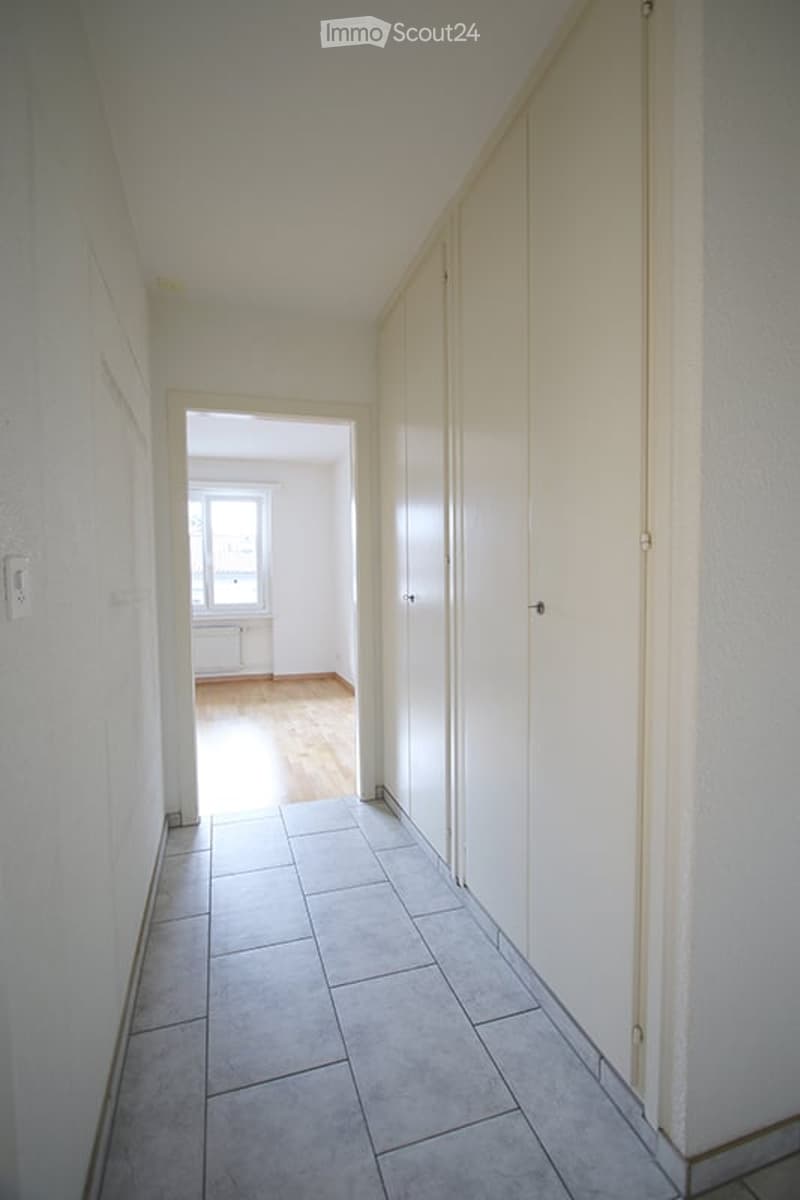 Empty hallway with tiled floor and wooden flooring visible in the adjacent room. White walls and closed doors.