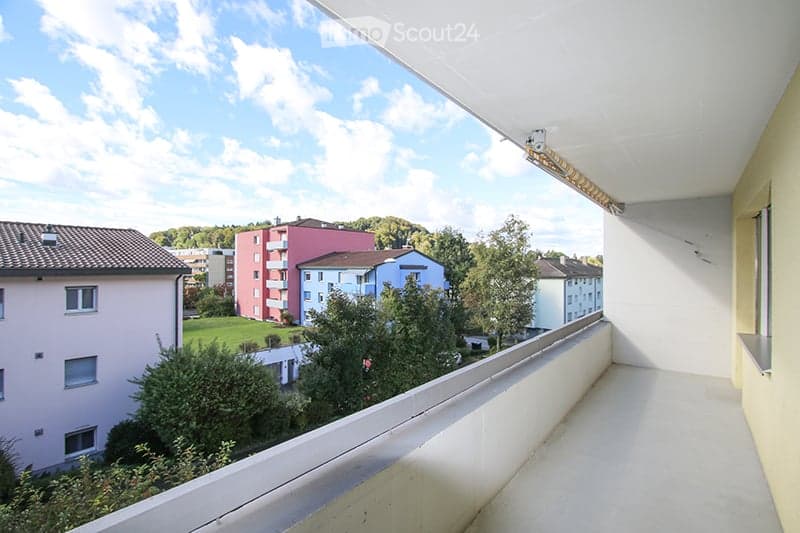Balcony with white railing, overlooking a residential area with buildings, trees, and a clear blue sky.