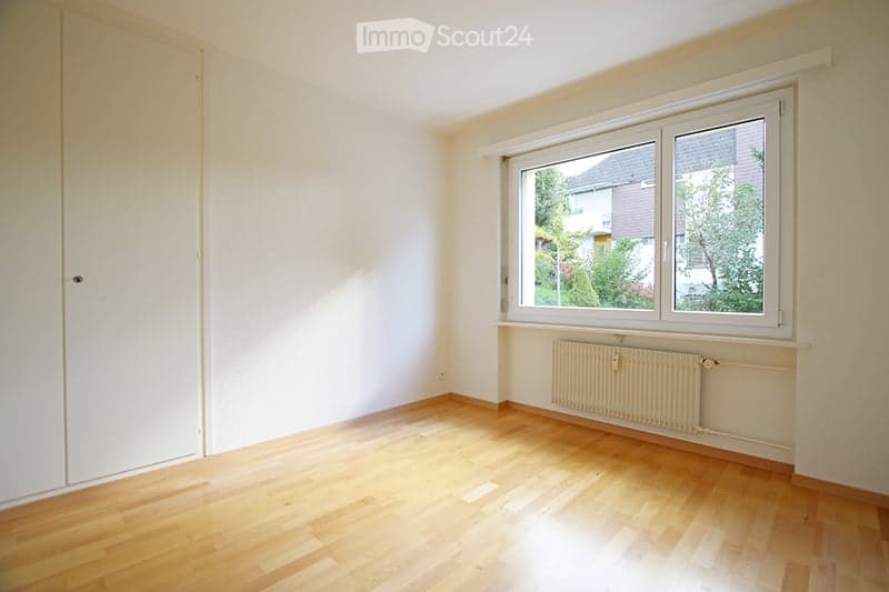 Empty room with wooden flooring, white walls, a large window with white frame, and a radiator below the window.