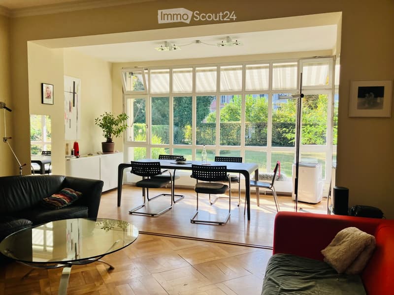 Bright living room with large windows, wooden flooring, a red sofa, a black leather sofa, a glass coffee table, a dining table with chairs, and a potted plant.