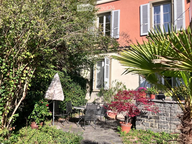 A lush garden area with a patio, outdoor furniture, potted plants, and a stone wall, adjacent to a building with shutters and windows.