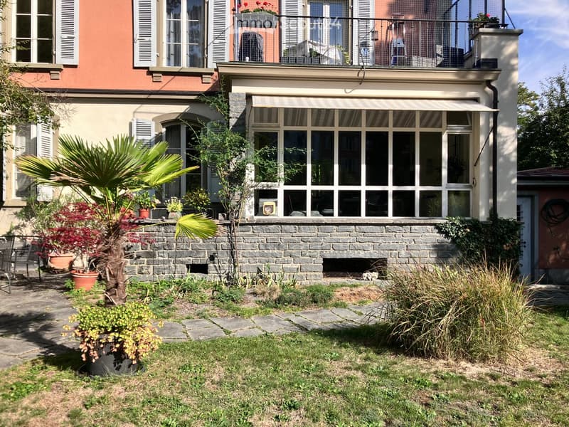 Garden area with stone pathway, potted plants, and a stone wall leading to a covered terrace with large windows and a balcony above.