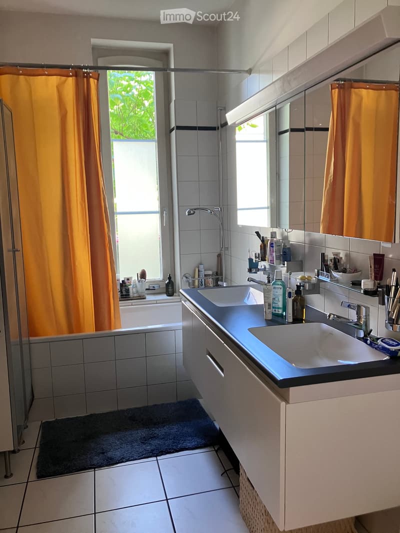 Modern bathroom with a double sink, mirror, bathtub, and shower area. Features include a dark countertop, white tiles, and a window with an orange curtain.