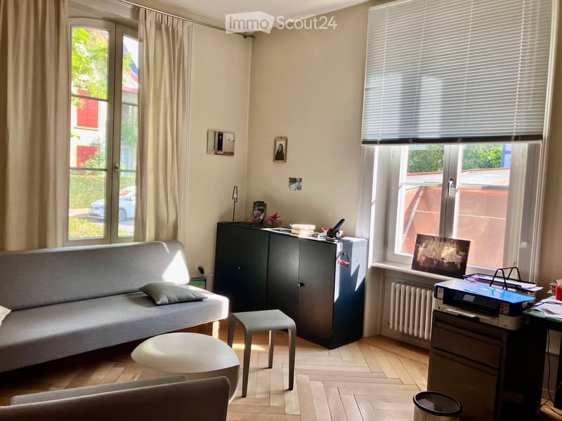 A well-lit living room with a grey sofa, a small round white stool, a grey chair, a dark cabinet, a small table with a laptop, a radiator, and a picture frame on the wall.