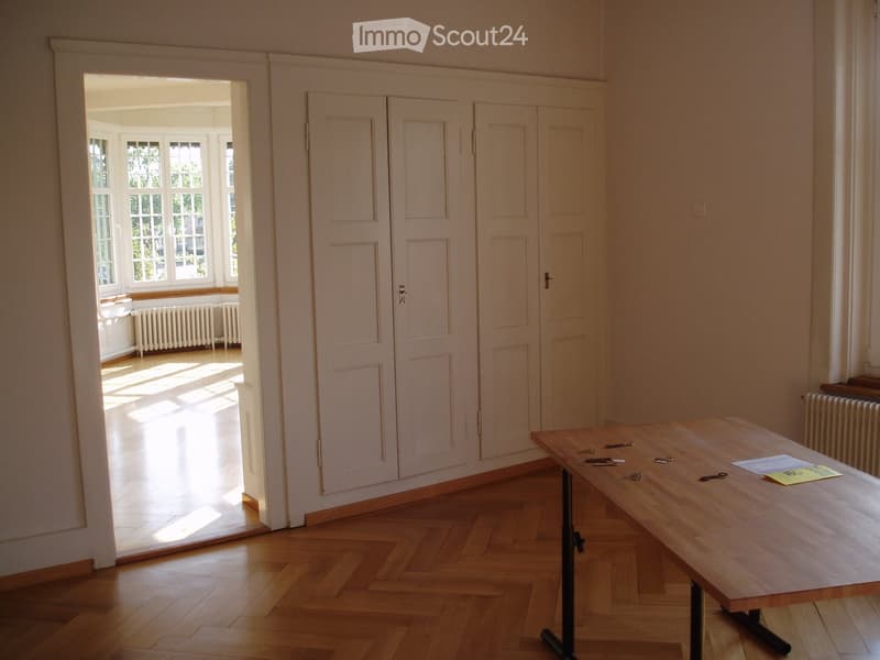 Bright room with wooden floor, large double doors, radiator, and a wooden table with keys and papers on it.