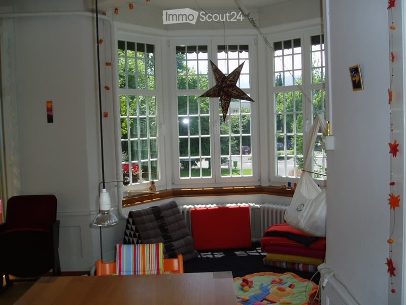 Brightly lit room with large windows, a sofa, chairs, a table, a hammock, and a colorful play mat with toys.