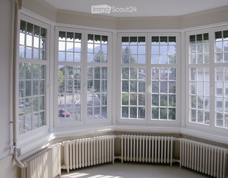 Empty room with large windows and radiators, offering a view of the street and surrounding area.