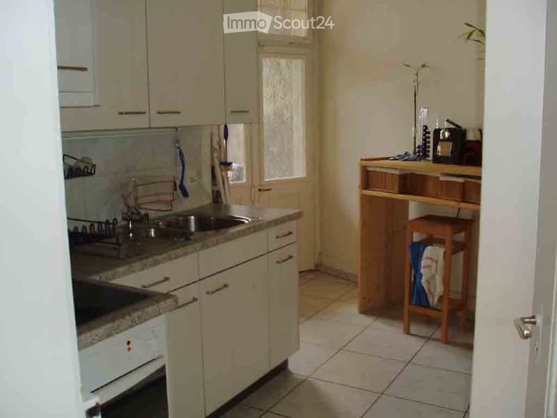 A small kitchen with light-colored cabinets, a granite countertop, a sink, stove, and oven. A small wooden counter with a stool and a vase is visible, along with a refrigerator in the background.