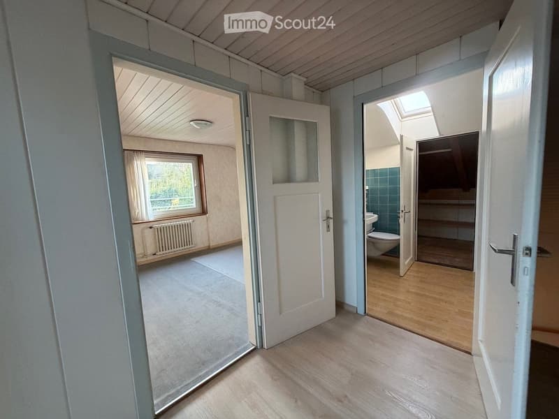 Empty hallway with wooden flooring, open doors leading to a room and a bathroom, visible window and radiator in the adjacent room.