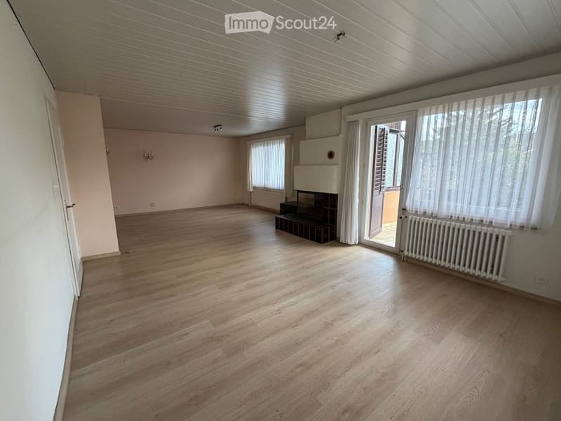 Empty room with wooden flooring, white walls, large windows with blinds, and a fireplace with a small bench in front of it.