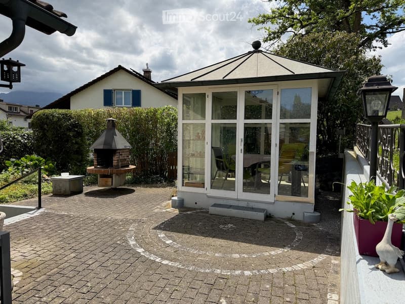 Garden area with a brick barbecue, a glass-enclosed gazebo, patio furniture, and a house with a white exterior and blue shutters.