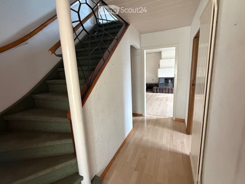 A well-lit hallway with wooden flooring, a staircase with a wooden handrail and metal balustrade, and an open doorway leading to another room.
