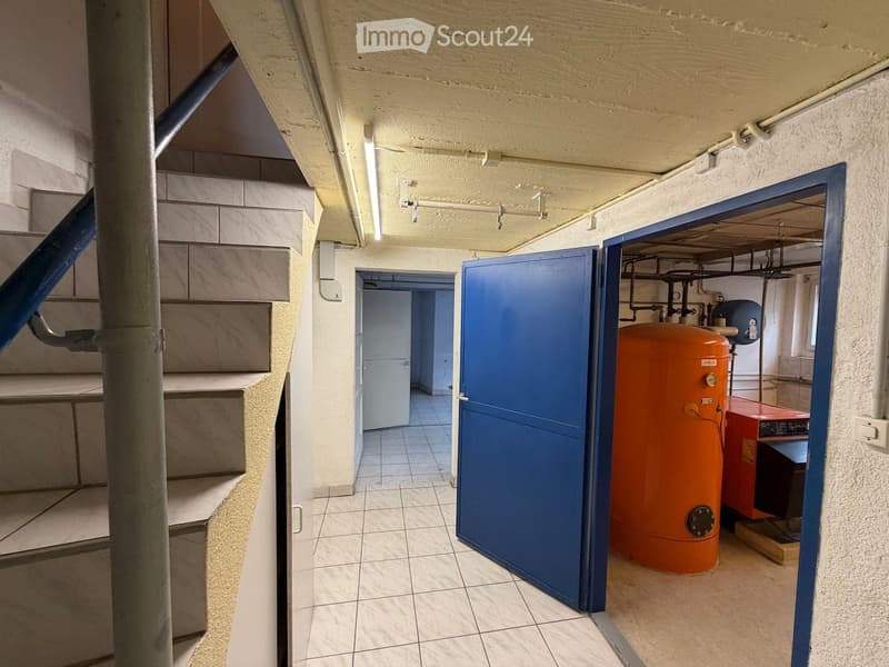 A narrow hallway with tiled floors, white walls, and a staircase leading upwards. An open door reveals a utility area with an orange water heater and other equipment.