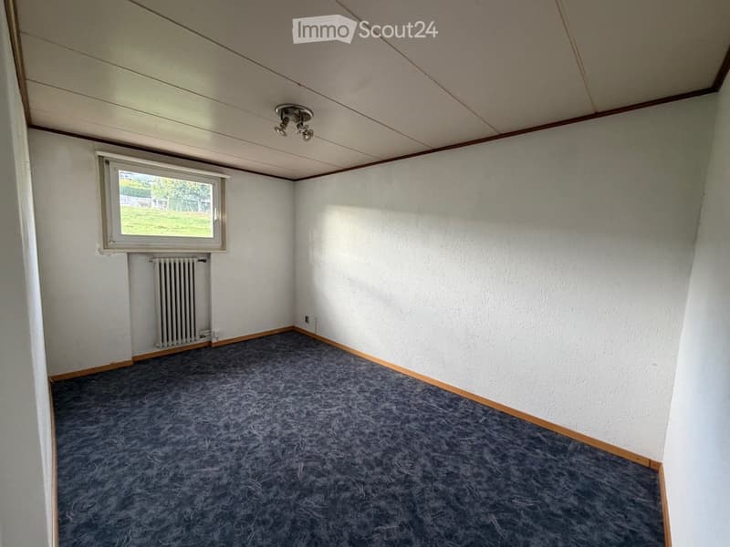 Empty room with grey carpet, white walls, wooden baseboards, a single window, radiator, and ceiling light fixture.