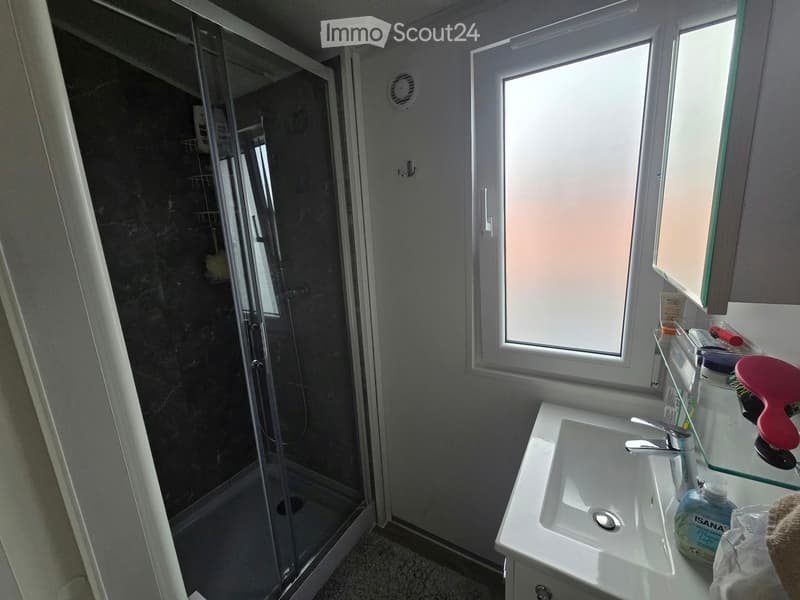 Modern bathroom with a white sink, chrome faucet, glass shelf, and a walk-in shower with dark tiles.