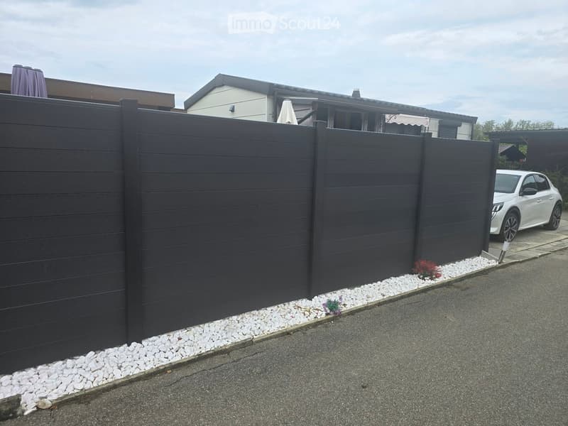 A modern dark grey wooden fence with white gravel border, white car parked nearby, and neighboring houses visible in the background.