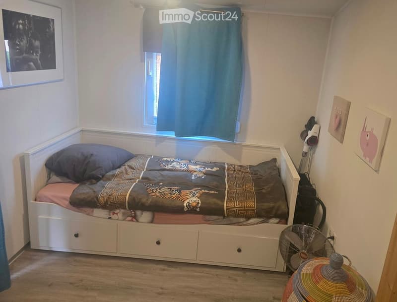 Cozy bedroom with a single bed, colorful bedding, a small fan, a round woven table, and a window with blue curtains.
