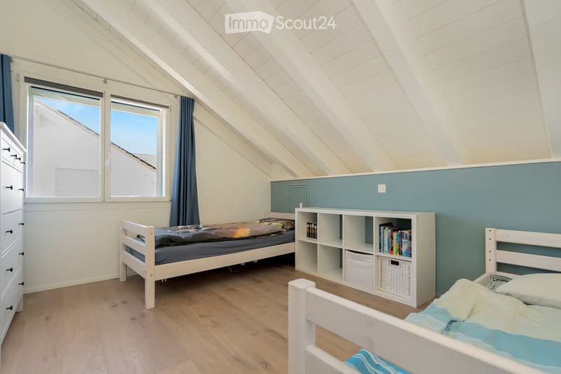 Bright attic bedroom with wooden floors, two single beds, a white bookshelf, and a tall window with blue curtains.