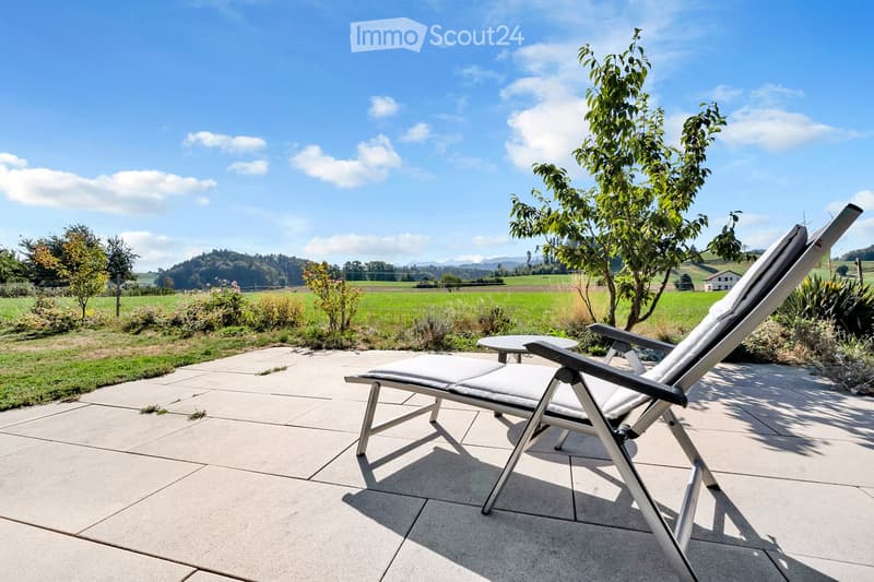 A sunny patio with a lounge chair and small table, surrounded by greenery and a scenic countryside view.