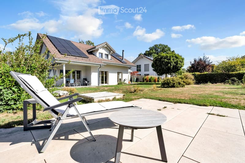 A modern patio area with a sun lounger and a round table, surrounded by a well-maintained lawn and a house with solar panels on the roof.