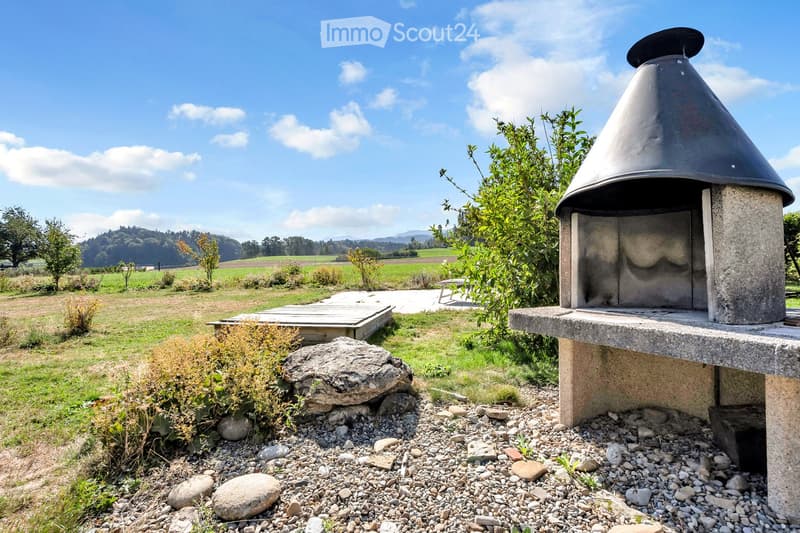 Outdoor area with a stone barbecue, wooden deck, scattered rocks, and lush greenery in a rural setting with open fields and distant hills.