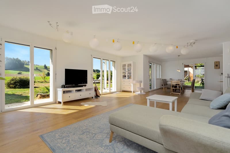 Bright living room with large windows and sliding doors leading to a garden, featuring a grey sofa, coffee table, TV, guitar, and a dining area with chairs and a table.