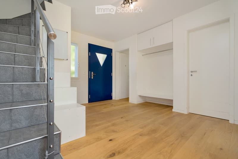 Bright hallway with wooden flooring, white walls, built-in closet, staircase with metal railing, and a blue door leading to another room.
