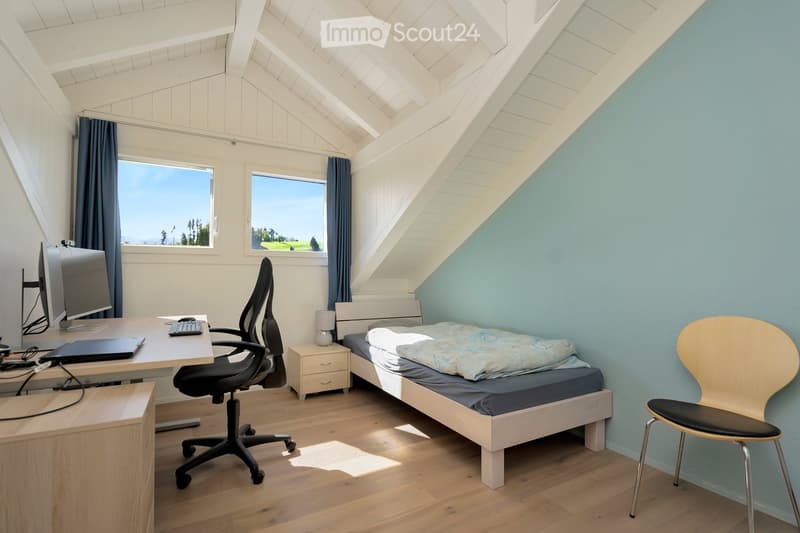 Bright bedroom with slanted wooden ceiling, large windows, bed, desk with chair, laptop, keyboard, mouse, and nightstand with lamp.