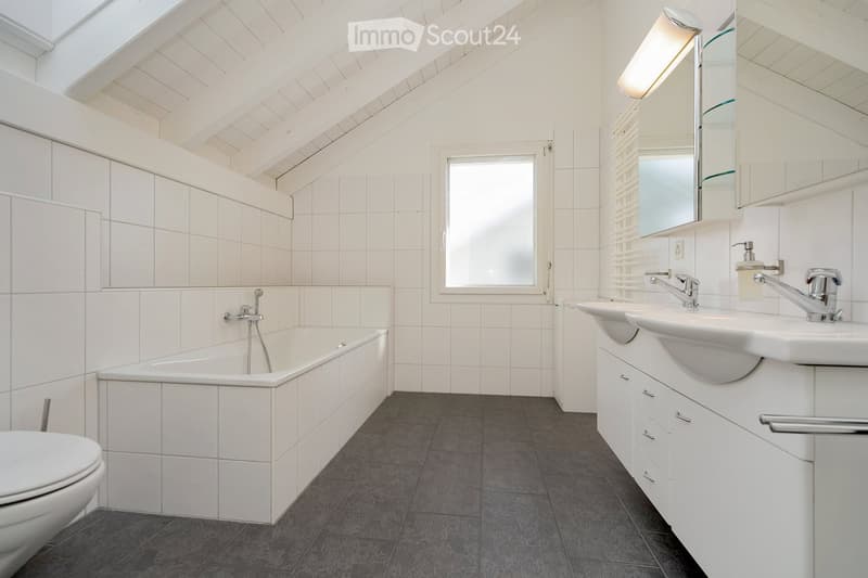 Modern bathroom with white tiles, a bathtub, toilet, double sink, and a large mirror. Features a skylight and a window with blinds.