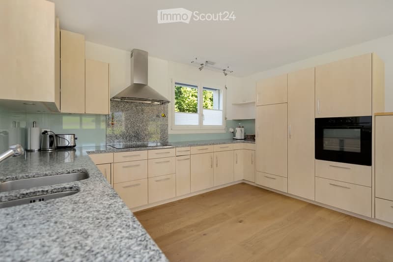 Modern kitchen with light-colored cabinets, granite countertops, and stainless steel appliances including a sink, oven, and range hood. The space features wooden flooring and large windows letting in natural light.