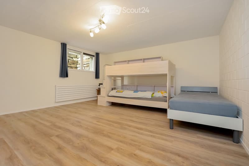 A small bedroom with wooden flooring, white walls, and a window with dark curtains. Features a bunk bed with built-in shelving and a single bed against the wall.