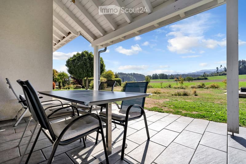 A covered patio with a table and chairs, overlooking a grassy field and distant hills.