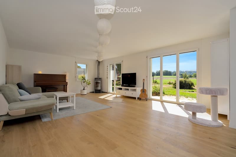Bright living room with wooden flooring, large windows, a grey sofa, a white coffee table, a piano, a guitar, a cat tree, and a television on a stand.