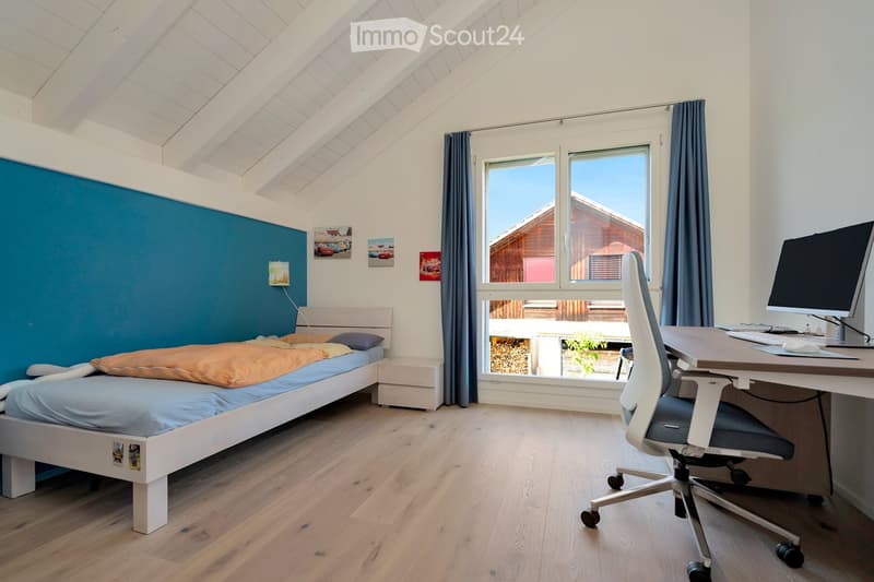 Bright bedroom with wooden flooring, slanted white ceiling, bed with blue and orange bedding, desk with computer, and large window with dark curtains.