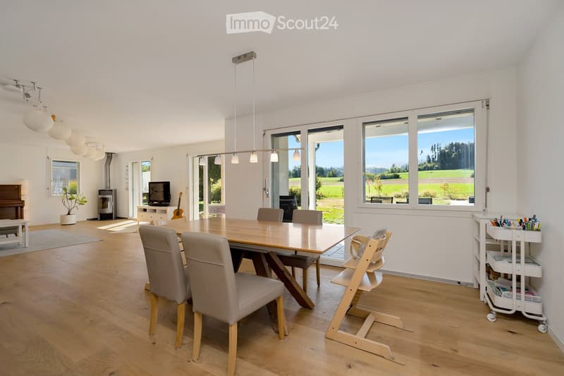 Bright, open dining area with wooden flooring, large windows overlooking a green landscape, a dining table with chairs, a baby chair, a piano, a guitar, a television, a potted plant, and a small cart with supplies.