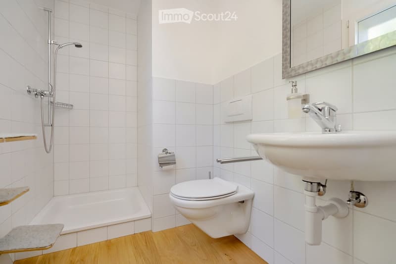 Modern bathroom with white tiled walls, wooden floor, bathtub, toilet, sink, mirror, and showerhead.