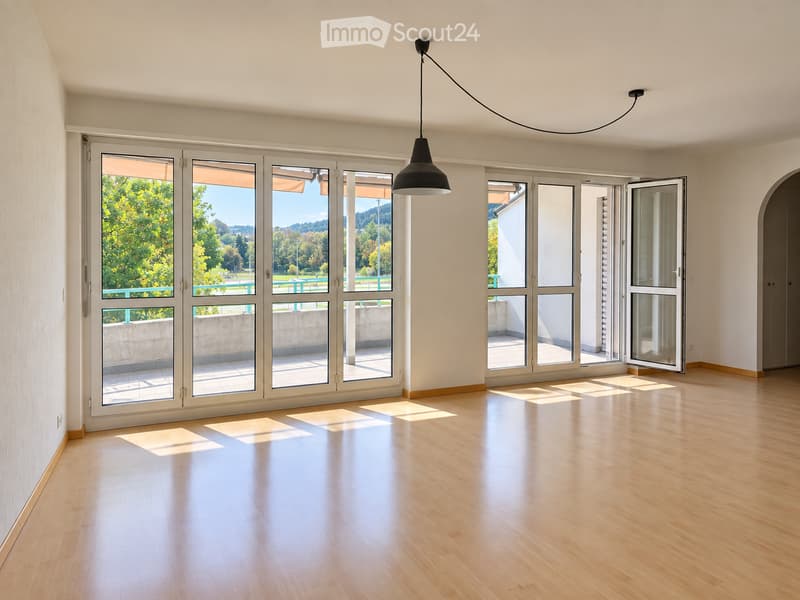 Empty room with light wooden flooring, large glass doors leading to a balcony, and a hanging pendant light fixture.