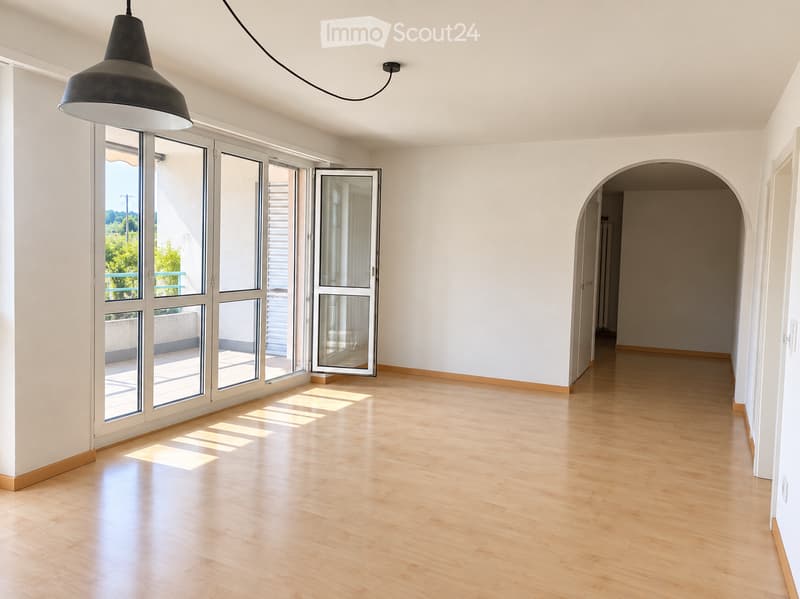 Empty room with light wooden flooring, white walls, large glass sliding doors leading to a balcony, and a hanging industrial-style lamp.