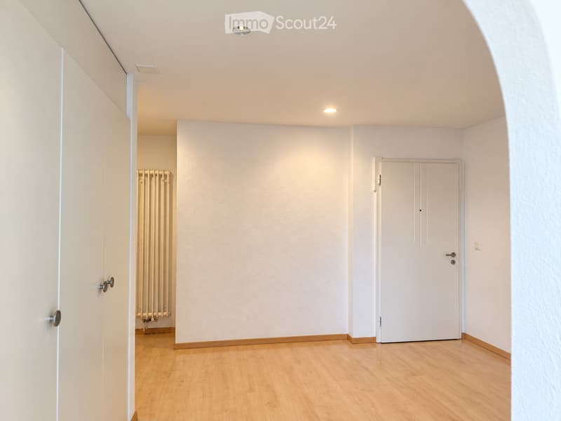 Empty room with light-colored walls, wooden flooring, and recessed ceiling lights. Features a radiator and two doors.